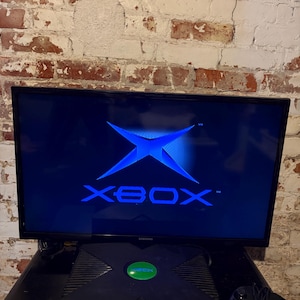 Könnte beinhalten: Eine schwarze Xbox-Konsole mit grünem Power-Knopf und schwarzem Controller. Der Fernsehbildschirm zeigt das Xbox-Logo in Blau. Die Konsole steht auf einer schwarzen Oberfläche vor einer Backsteinmauer.