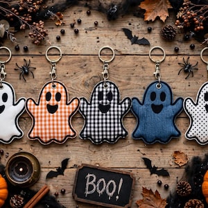May include: Five ghost-shaped keychains with metal rings and clasps. The ghosts feature different patterns: solid white, orange gingham, black and white buffalo check, solid blue denim, and white with black polka dots, all with black eyes and mouths.