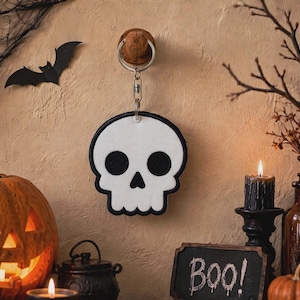 May include: A white skull-shaped keychain with a black border and black eye and nose cutouts, attached to a silver metal split ring. The skull features a smooth, flat surface and is suspended from a circular wooden wall hook.