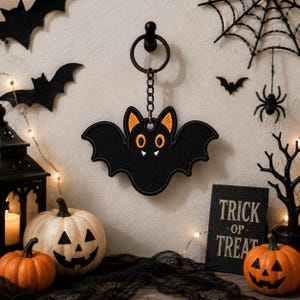 May include: A black bat-shaped keychain featuring embroidered orange inner ears, orange oval eyes, and two small white fangs. The bat is attached to a dark metal split ring by a short chain, showcasing detailed stitching throughout the design.