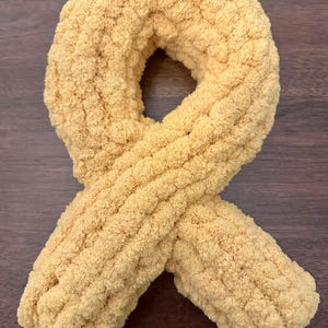 May include: A mustard yellow awareness ribbon crafted from thick, plush, chenille-style yarn. The soft, textured material is looped into a classic ribbon shape with two overlapping ends at the bottom, creating a continuous, rounded, and tactile form.
