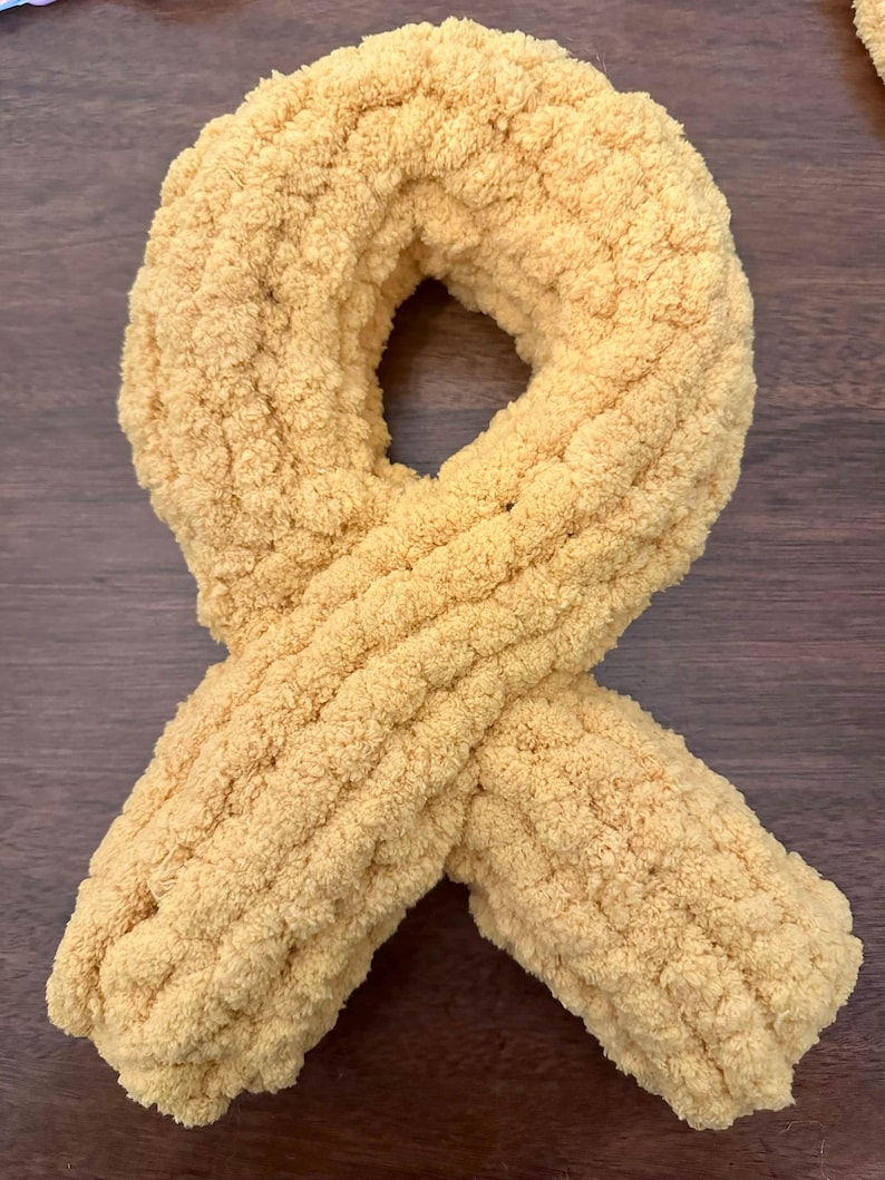 May include: A mustard yellow awareness ribbon crafted from thick, plush, chenille-style yarn. The soft, textured material is looped into a classic ribbon shape with two overlapping ends at the bottom, creating a continuous, rounded, and tactile form.