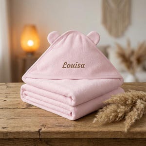 Könnte beinhalten: Zwei rosa Babyhandtücher, gestapelt auf einer Holzoberfläche. Das obere Handtuch ist mit einer Kapuze und kleinen Ohren gefaltet und trägt den Namen "Louisa" in brauner Stickerei.