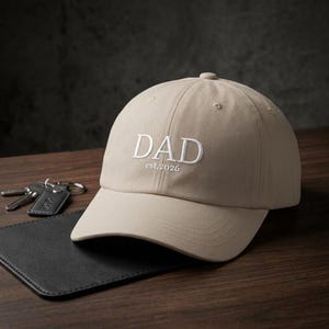 Könnte beinhalten: Beige Baseballcap mit dem weißen gestickten Wort "DAD" und dem Text "est.2026". Die Kappe liegt auf einer Holzoberfläche neben einer schwarzen Brieftasche und Schlüsseln.