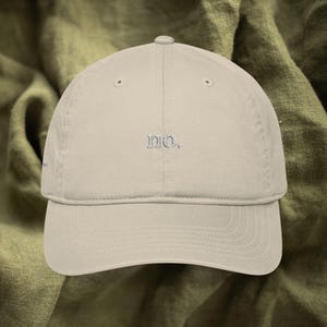 Könnte beinhalten: Beige Baseballkappe mit gebogenem Schirm. Das Wort "no." ist in hellblauem Faden auf die Vorderseite gestickt. Die Kappe ist aus weichem Stoff und hat einen klassischen, lässigen Stil.