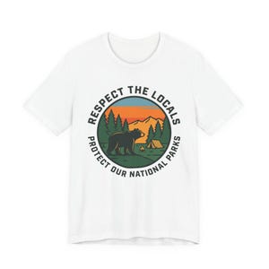 Könnte beinhalten: Weißes T-Shirt mit einem kreisförmigen Grafikdesign, das einen Bären, Bäume, Berge und ein Zelt zeigt. Der Text lautet "Respect the Locals, Protect Our National Parks."