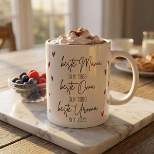 Könnte beinhalten: Weiße Keramiktasse mit den Worten "beste Mama, beste Oma, beste Uroma" in schwarzer Schrift, zusammen mit Daten. Die Tasse ist mit einem Getränk mit Schlagsahne und roten Herzakzenten gefüllt. Die Tasse steht auf einem Marmortablett.