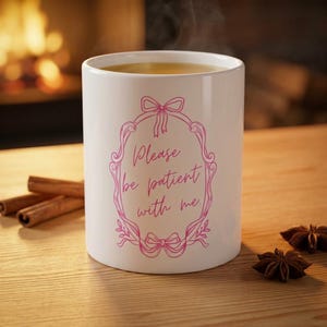 Könnte beinhalten: Weißer Keramikbecher mit rosa floralem Design und dem Text "Please be patient with me." Der Becher enthält ein warmes Getränk und ist von Zimtstangen und Sternanis umgeben.