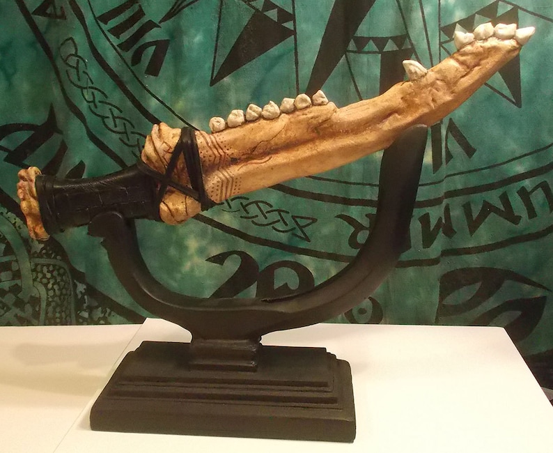 May include: A decorative bone-like dagger with a black leather wrap and silver teeth, displayed on a black stand.