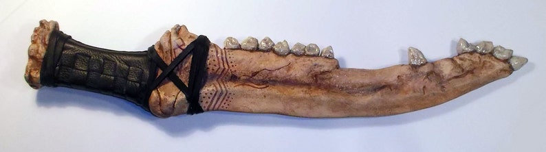 May include: A bone-like dagger with a black leather handle and a row of teeth along the blade. The handle is wrapped in black leather and has a decorative pattern.