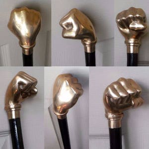 May include: A black walking cane features a handle shaped like a brass-coloured fist. The handle has a metallic sheen, and the cane has a brass collar. A classic accessory.