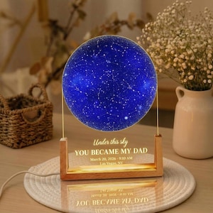 May include: A wooden-framed night light with a blue, starry sky design. The text "Under this sky YOU BECAME MY DAD" is displayed, along with a date and time. The light sits on a white placemat, with a vase of flowers in the background.