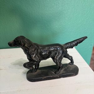 Puede incluir: Figura de perro de hierro fundido negro en posición de caminar. El perro tiene una cola larga y está montado sobre una base rectangular. La figura está colocada sobre una superficie blanca y una pared verde azulado.
