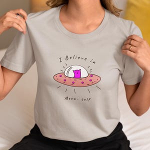 May include: A light grey short-sleeved t-shirt featuring a graphic of a pink cat inside a flying saucer. The text above the saucer reads I Believe in and the text below reads Meow- self. The saucer is pink with small purple circular accents.