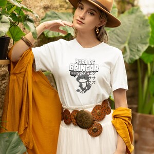May include: A white short-sleeve t-shirt featuring a graphic of a grey and white striped cat with the text "HORA DE BRINCAR" above it and Japanese characters to the side. The shirt is paired with a wide brown leather belt with circular metal accents.