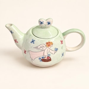 May include: A light green ceramic teapot featuring a hand-painted illustration of an angel with orange hair and white wings. The angel holds a blue heart and carries a brown basket. Small blue and red flowers decorate the teapot and lid.