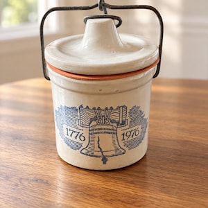 Pode incluir: Um pote de cerâmica na cor creme com tampa correspondente e uma alça de arame de metal escuro. A frente apresenta um gráfico azul da Liberty Bell com as datas 1776 e 1976 impressas em cada lado. A tampa tem um botão central e uma borda laranja.