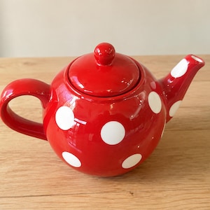 Può includere: Una teiera rotonda in ceramica rossa lucida caratterizzata da un motivo a grandi pois bianchi. La teiera include un coperchio rosso abbinato con un piccolo pomello, un beccuccio curvo e una robusta impugnatura laterale, con finitura smaltata.