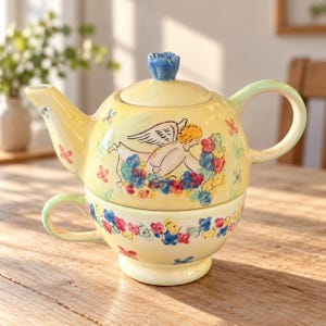 May include: A yellow ceramic tea-for-one set featuring a teapot stacked on a teacup. The teapot has a blue flower-shaped lid handle and a painted angel surrounded by colourful flowers. The teacup base is decorated with a matching floral pattern.