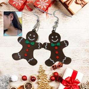 May include: A pair of brown gingerbread man earrings with silver-tone fishhook wires. Each gingerbread figure features a green bow tie, three red circular buttons, and white wavy icing details on the arms and legs. The earrings are shown hanging against a light wood background with holiday decorations.
