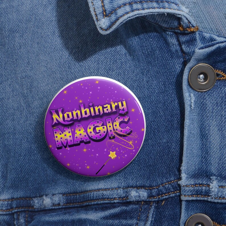 May include: A round, purple button with the words "Nonbinary MAGIC" in yellow and purple lettering. The button has a starry background and a small wand graphic. The button is pinned to a denim jacket.