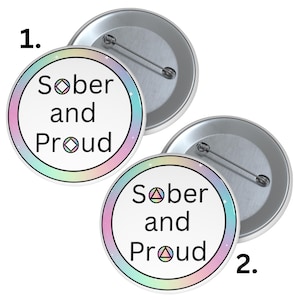 May include: Two round buttons with the text "Sober and Proud" in black. The buttons have a white background with a pastel rainbow border and a silver pin back. The buttons feature the Alcoholics Anonymous symbol.