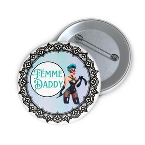 May include: A round button with a graphic of a person in black lingerie, holding a whip. The text "Femme Daddy" is in a teal font, surrounded by a black lace design and a light blue and white background.