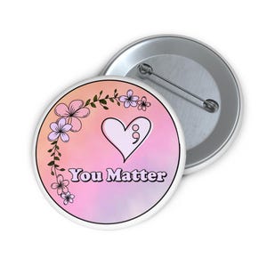 Könnte beinhalten: Ein runder Button mit einem rosa-pfirsichfarbenen Farbverlauf im Hintergrund. Der Button zeigt ein weißes Herz mit einem Semikolon, floralen Akzenten und den Worten "You Matter" in einer Schriftart. Ein zweiter Button ist im Hintergrund sichtbar.