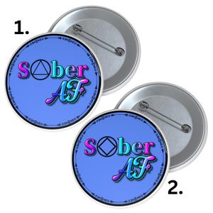 May include: Two round, metal pinback buttons with a light blue background. The buttons feature the text "Sober AF" in a gradient of pink, blue, and teal, with a black triangle and diamond symbol. The buttons have a decorative border.