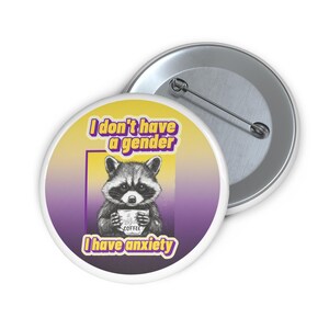 Può includere: Un bottone rotondo con un'illustrazione di un procione che tiene una tazza di caffè. Il bottone ha il testo "I don't have a gender, I have anxiety" in giallo e viola. Un secondo bottone è visibile sullo sfondo.
