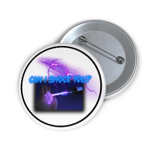 May include: A white button with the text "CAN I SHOCK YOU?" in blue neon lettering. The button features a graphic of a hand holding a device with a lightning bolt design. A second button is visible.