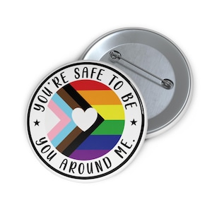 Puede incluir: Un botón redondo blanco con el texto "YOU'RE SAFE TO BE YOU AROUND ME." El botón presenta un diseño de bandera arcoíris con un corazón blanco y un borde negro. Un segundo botón es visible en el fondo.