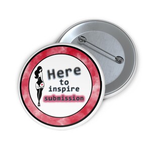 Könnte beinhalten: Ein weißer Ansteckbutton mit rotem Rand und dem Text "Here to inspire submission". Der Button zeigt eine Grafik einer Frau in einem Korsett. Ein zweiter Button ist im Hintergrund sichtbar.