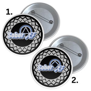 May include: Two round pinback buttons with a silver backing. The buttons have a black center with the words "Sober AF" in blue and white, surrounded by a gray geometric pattern. A blue and white triangle and square symbol is in the center.