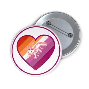 May include: A white button pin with a heart-shaped design in the colors of the lesbian pride flag. The heart contains an axolotl with heart-shaped eyes and floral accents. A second button pin is visible in the background.