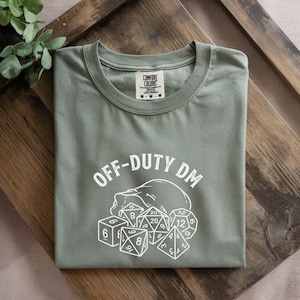 May include: Sage green t-shirt with the text "OFF-DUTY DM" above a skull and dice graphic. The dice include a six-sided die, an eight-sided die, a twenty-sided die, and a twelve-sided die. The shirt is folded on a wooden surface.
