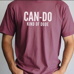 May include: A mauve short-sleeved t-shirt featuring the white text CAN-DO KIND OF DUDE printed on the chest. The shirt has a crew neckline and a standard fit, with the text positioned centrally across the upper torso area of the garment.