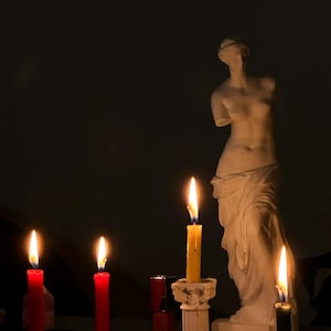 Op de afbeelding: Een donkere scène met een klassiek wit beeld en verschillende brandende kaarsen. Drie rode kaarsen en een gele kaars branden fel, samen met een groene kaars. De kaarsen staan in verschillende houders, wat een warme sfeer creëert.