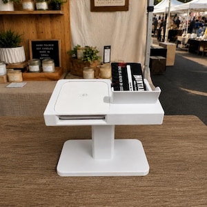 May include: A white point-of-sale terminal stand featuring a flat square payment reader surface with a contactless symbol and an integrated side slot holding a black card with the text MONTANA and GIVE US A REVIEW ON GOOGLE.