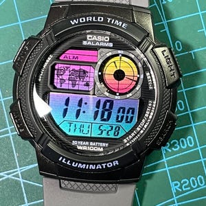 May include: A black Casio digital watch with a multi-colored gradient display featuring a world map, a circular analog-style dial, and digital time readout showing 11:18, date THU 5-28, 10 YEAR BATTERY, WR100M, and ILLUMINATOR text on the bezel.