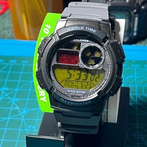 May include: A black and gray Casio digital watch with a yellow display showing the time 5:33:02. The watch has the text "WORLD TIME" and "ILLUMINATOR". The watch is on a black stand with a green tag that says "10".