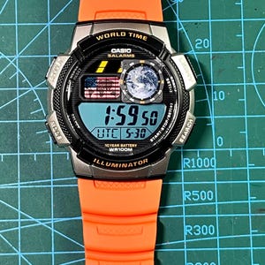 May include: A Casio digital watch with a black and silver face, orange band, and a digital display showing the time. The watch face includes the text "WORLD TIME", "CASIO SALARMS", and "ILLUMINATOR". The watch also features a small American flag graphic.