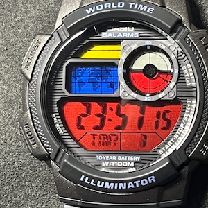 May include: A Casio World Time digital watch with a grey resin strap and case. The face features a red digital display, a blue world map graphic, a yellow accent bar, and a circular sub-dial with red and white segments. Text includes WORLD TIME, 5 ALARMS, 10 YEAR BATTERY, WR100M, and ILLUMINATOR.