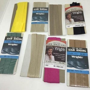 Könnte beinhalten: Verschiedene Nahtband- und Schrägband-Packungen der Marke Wrights in Farben wie Gelb, Beige, Schwarz und Pink. Die Verpackungen zeigen Produktinformationen und Bilder. Auf den Packungen steht "Soft and Easy Seam Binding" und "Wide Single Fold Bias Tape."