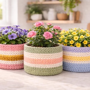 Op de afbeelding: Drie kleurrijke gehaakte bloempotten gevuld met bloemen. De potten zijn cilindrisch met horizontale strepen in pastelkleuren: roze, geel, wit, groen en blauw. De bloemen zijn onder meer paarse petunia's, roze rozen en gele calibrachoa.