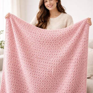 May include: A light pink crocheted blanket with a textured pattern. The blanket is held up, showcasing its size and design. The soft, pastel color and open weave suggest a cozy, handmade item, perfect for home decor.