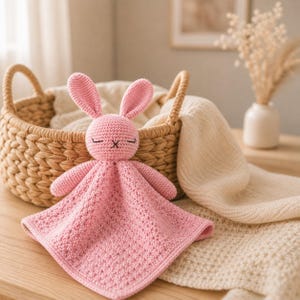 May include: A handmade pink crochet bunny security blanket with closed eyes and an X for a mouth. The bunny is attached to a square blanket. The blanket is on a wooden table near a wicker basket and a cream-colored blanket.