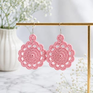 May include: A pair of pink crochet earrings with a circular design and scalloped edges. The earrings are displayed on a gold-colored earring stand. The background includes a white vase with white flowers and a marble surface.