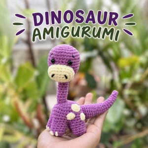 Op de afbeelding: Een handgemaakte paarse amigurumi dinosaurus met een lange nek, crèmekleurige snuit en witte vlekken. De tekst "DINOSAUR AMIGURUMI" wordt boven de dinosaurus weergegeven. De dinosaurus wordt in een hand gehouden tegen een vage groene achtergrond.