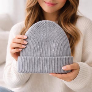 May include: A light gray ribbed knit beanie hat with a folded brim. The hat is made of a soft, warm material and is a classic winter accessory. The beanie is held in the hands.